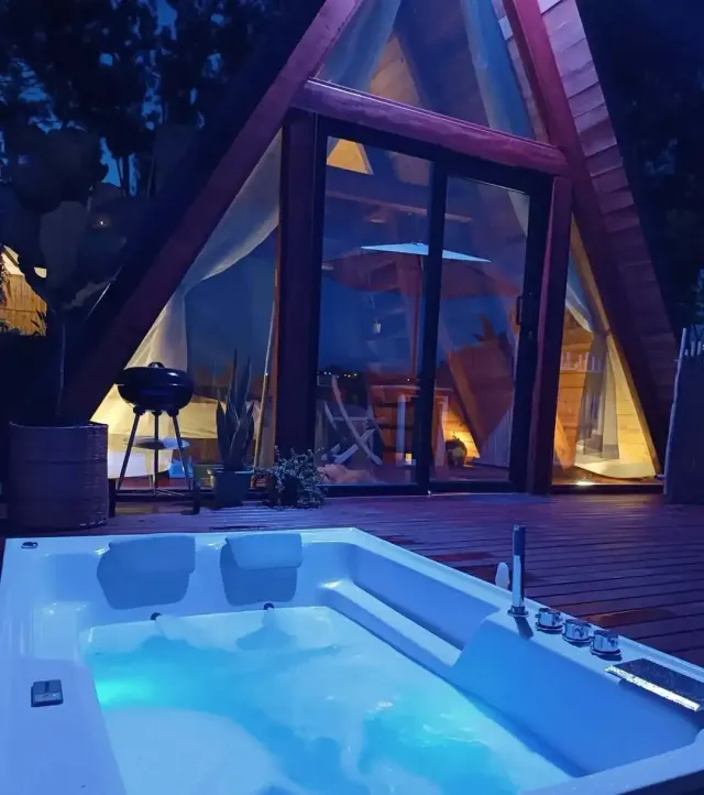 Glamping nocturno con cabaña en madera y jacuzzi privado al aire libre, iluminación cálida y entorno natural ideal para descansar.