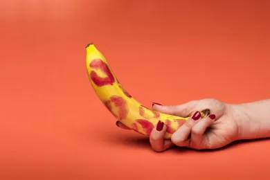 Mano con uñas rojas sostiene banana con marcas de labial, concepto creativo de juegos en pareja y diversión íntima