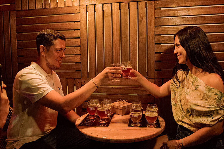 Pareja brindando con cervezas artesanales en un espacio íntimo y acogedor, con una mesa de madera decorada con detalles como una caja sorpre