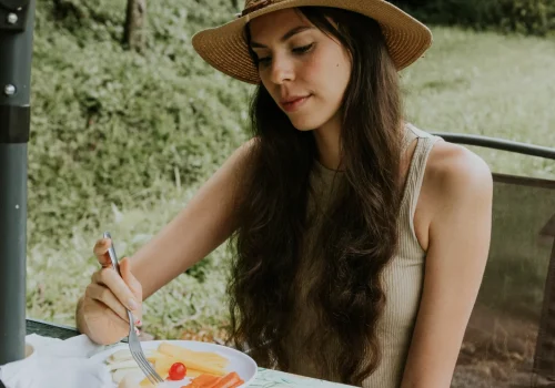 La imagen muestra una mujer disfrutando de una experiencia gastronómica al aire libre, degustando un café junto a un plato de frutas frescas. Rodeada por un entorno natural con árboles y vegetación, la escena resalta la conexión con la naturaleza mientras saborea una comida ligera. Ideal para promocionar actividades de turismo gastronómico, experiencias de cata de café o eventos al aire libre.