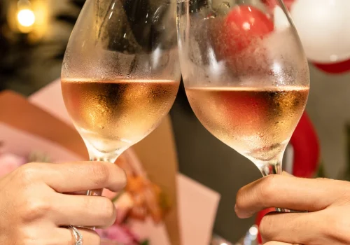 Dos copas de vino rosado son alzadas en un brindis, con un fondo decorado con globos y arreglos florales, creando un ambiente romántico y elegante. Esta imagen es ideal para promocionar eventos de cata de vinos, celebraciones especiales o experiencias románticas en lugares exclusivos.