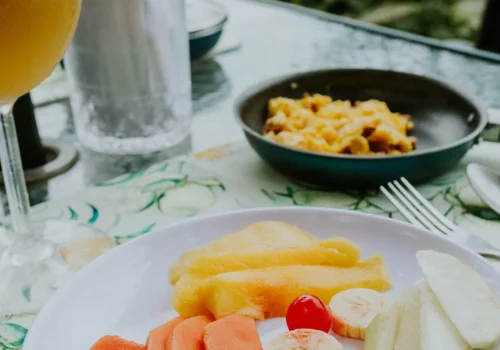 Un desayuno colorido al aire libre compuesto por frutas frescas, huevos revueltos y una bebida espumosa, con un paisaje natural al fondo. Perfecto para destacar servicios de desayunos sorpresa, brunches al aire libre o escapadas gastronómicas en entornos rurales.