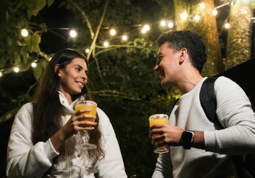 La imagen muestra a una pareja disfrutando de una bebida al aire libre bajo luces colgantes en un entorno nocturno rodeado de árboles. Ambos lucen relajados y felices, destacando un ambiente acogedor y mágico. Perfecta para promocionar experiencias de picnic romántico, actividades al aire libre o turismo sostenible.