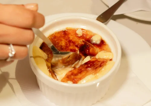 La imagen muestra un delicioso crème brûlée servido en un recipiente blanco, siendo degustado con una cuchara. El caramelo dorado en la parte superior destaca la textura crujiente, mientras que la crema suave y los acompañamientos resaltan el detalle gourmet del postre. Ideal para promocionar restaurantes, eventos gastronómicos, o servicios de catering especializados en repostería de alta calidad.