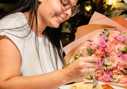 Una mujer sonriente degusta un plato de pasta en un restaurante al aire libre, rodeada de una decoración elegante con luces cálidas y flores. La escena combina lo gourmet con un entorno acogedor, ideal para destacar rutas gastronómicas, experiencias culinarias o destinos turísticos enfocados en la comida.